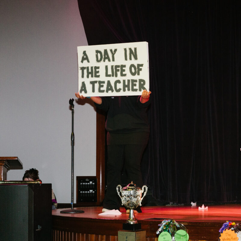 individual on a stage holding up a sign that says "a day in the life of a teacher"