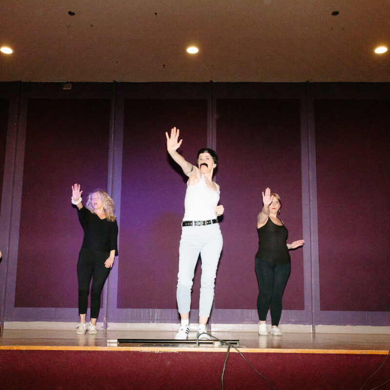 one performer wearing a white outfit and a mustache with four performers in black outfits holding one hand up on the stage