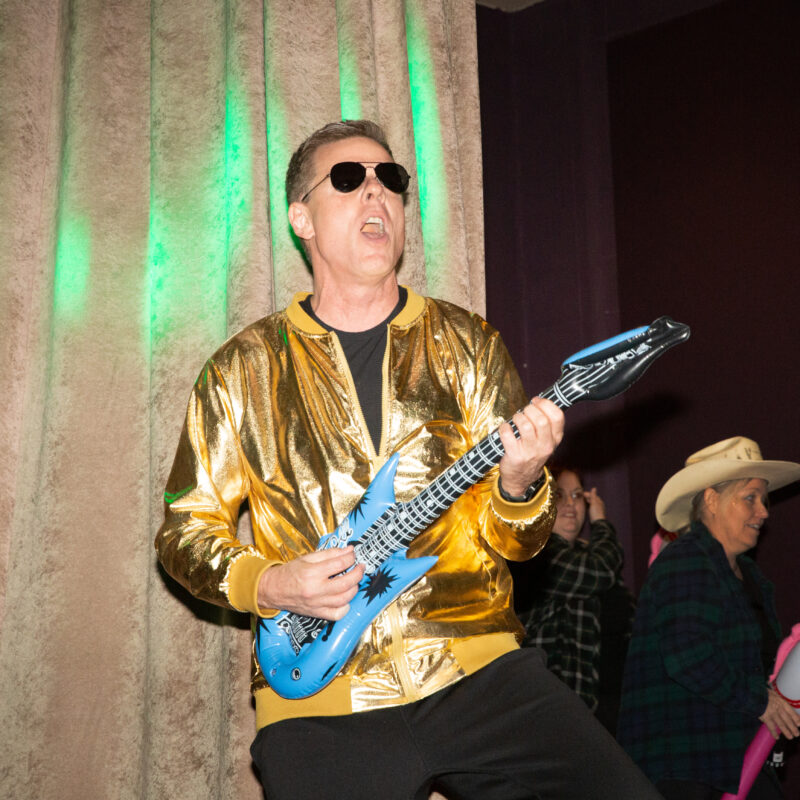 man wearing sunglasses and a gold jacket pretending to play an inflatable guitar