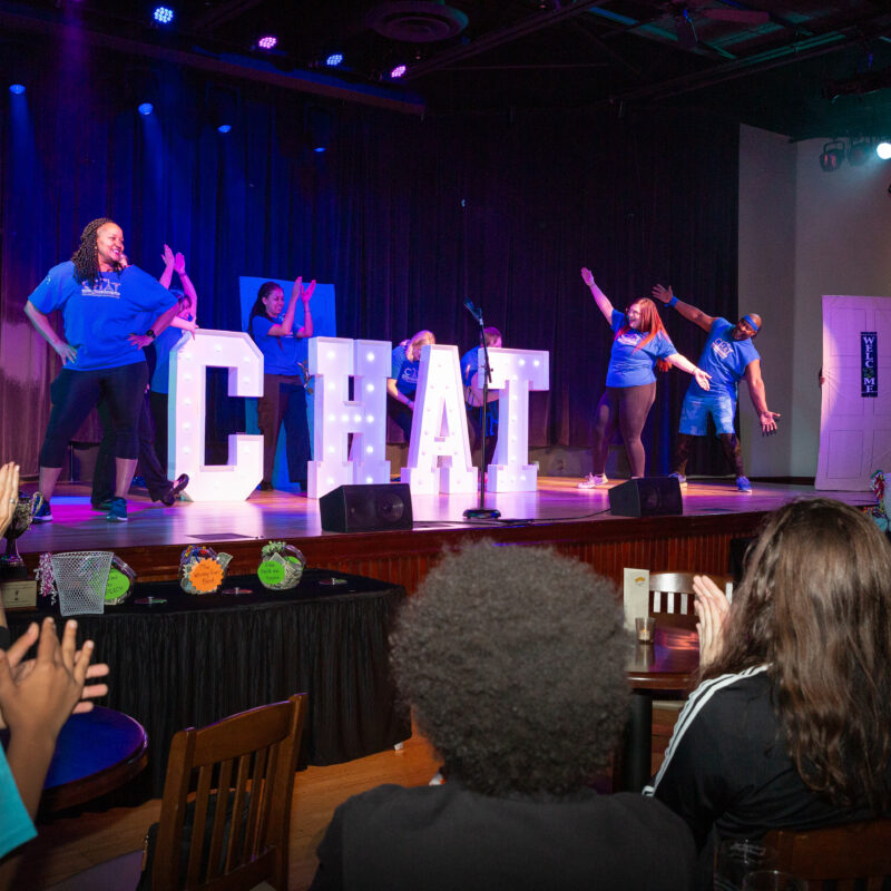 six performers on a stage posing next to large light up letters spelling out chat