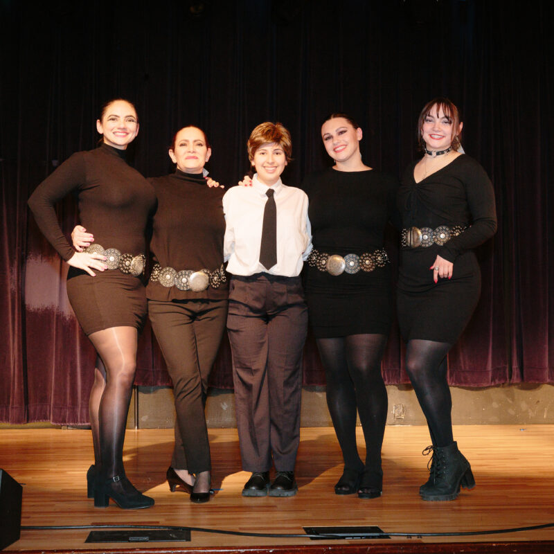 one person in a formal outfit and four women in black dresses posing on a stage