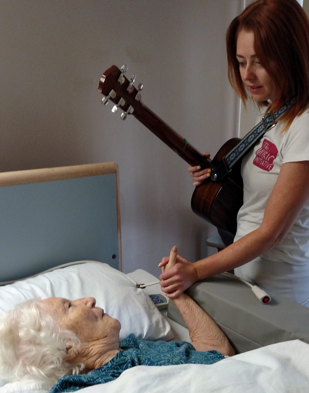 older woman in a hospital bed with a younger woman attending her holding a guitar